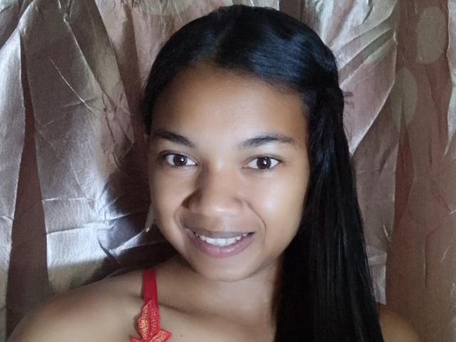 MayahMagique has smooth brown skin, black straight hair, and a cute smile showing white teeth. She's wearing a red lace bra, and her brown eyes are looking forward.