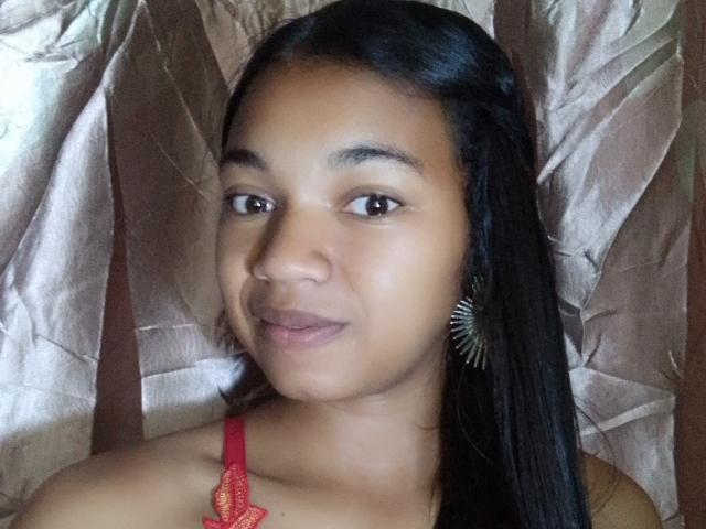 MayahMagique with brown skin, long black hair, and small red lace top, wearing a spiky earring, smiling softly at the camera.