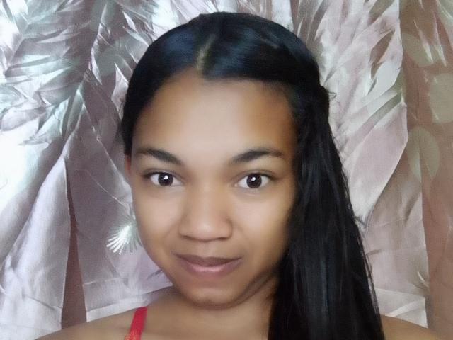 MayahMagique has smooth brown skin, long black hair, brown eyes, and a subtle smile. She's wearing a red top and has a small white flower earring on her right side.