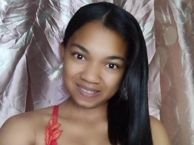 MayahMagique with dark brown skin, straight black hair, and small nose, smiling, wearing red lace top, and large fluffy earrings, showing her cute face and medium-sized tits.