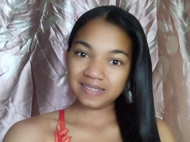 MayahMagique has dark brown skin, straight black hair, brown eyes, small nose, and is smiling with red lace top showing her tits. She's wearing feather earrings.