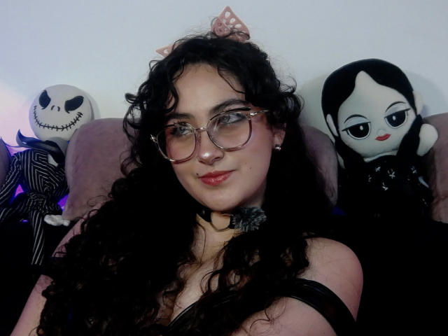 HannaCeleste with curly black hair, glasses, and black choker, sitting between Jack Skellington and a black-eyed doll, looking cute with pink bow on head. Boobs visible in black top.