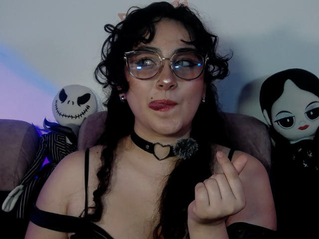 HannaCeleste with curly black hair, glasses, and a black choker with a heart, pointing with her index finger, wearing a black top, sitting between Jack Skellington and Sally.