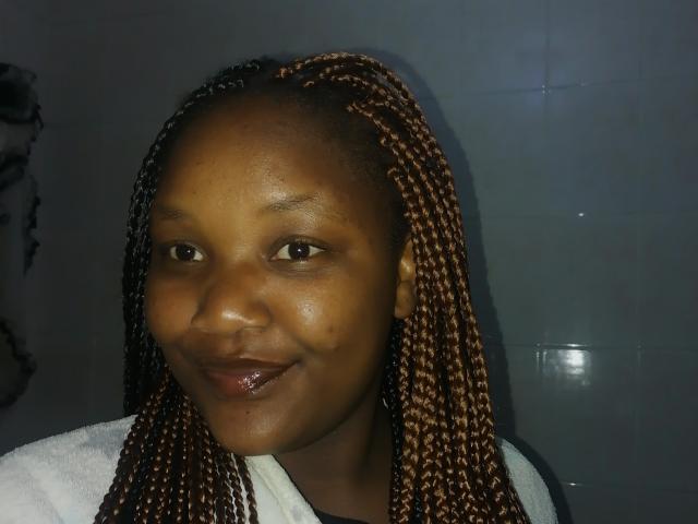 QueziQueen with dark brown skin, small nose, and brown braided hair, smiling with glossy lips, wearing a white towel. She has a slight pimple on her right cheek.