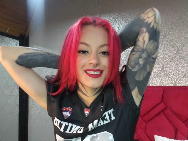 DahianaKlimt with bright red hair, black tattooed arms, nose ring, and red lipstick, smiling, wearing a black football jersey, arms raised. Boobs not visible.