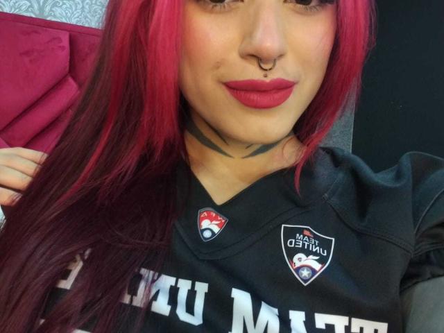 DahianaKlimt has red and black hair, a nose ring, and dark lipstick. She's wearing a black sports shirt with NFL logos, showing tattoos and a confident smile.