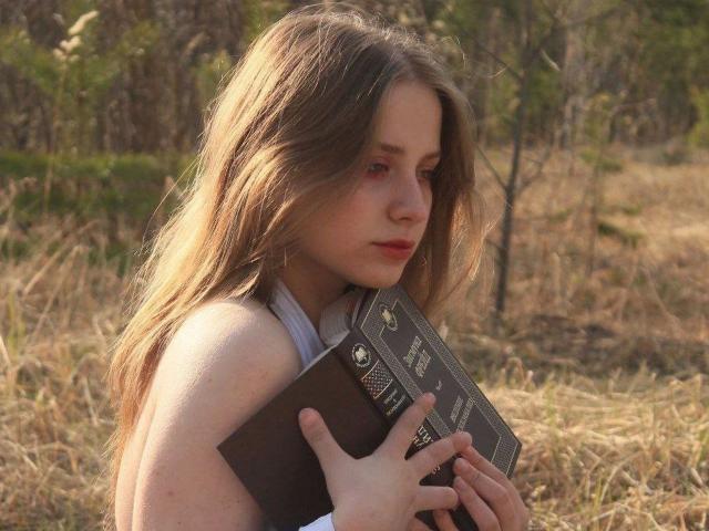 Annie, con cabello largo y ondulado de color marrón, sosteniendo un libro, mirando hacia abajo con una expresión ligeramente triste, vestida con un top blanco de tirantes, en un campo soleado y cubierto de hierba..
