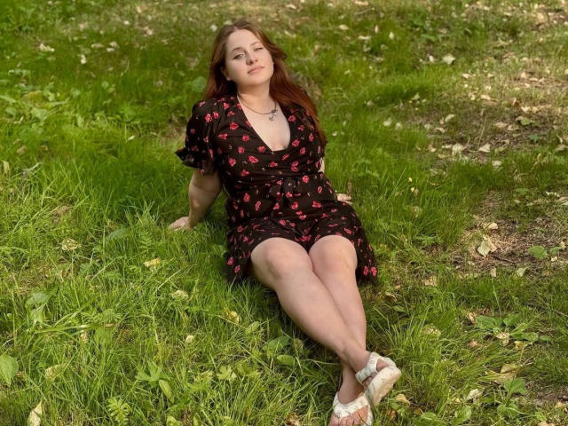 AnnieWow – schlank, mit heller Haut und roten Haaren – sitzt auf dem Gras, trägt ein schwarzes Kleid mit rosa Mustern, das ihren Hintern und ihre Beine zeigt, weiße Sandalen und wirkt entspannt..