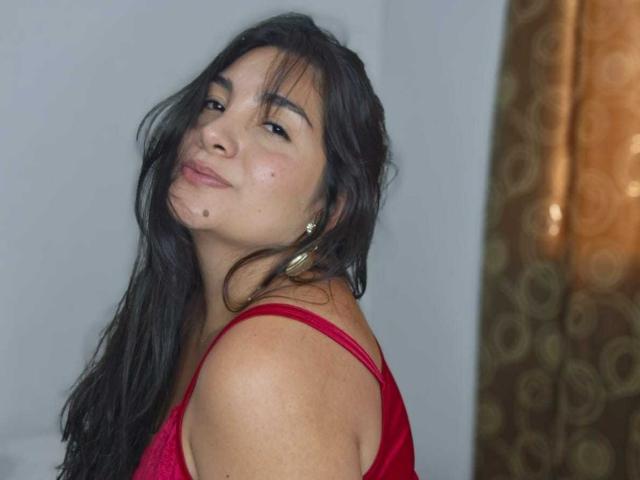 CherrySweetX with long black hair, red dress, small tits, and a cute mole on her cheek, smiling slightly with a relaxed, sexy vibe, wearing gold hoop earrings.