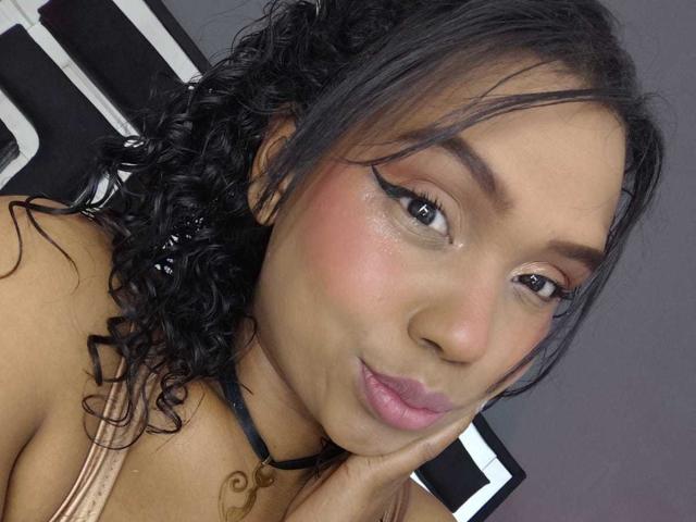 SucejPonce has curly black hair, brown skin, and pink lips. She's wearing black eyeliner and a black choker, with her hand touching her cheek, looking cute.