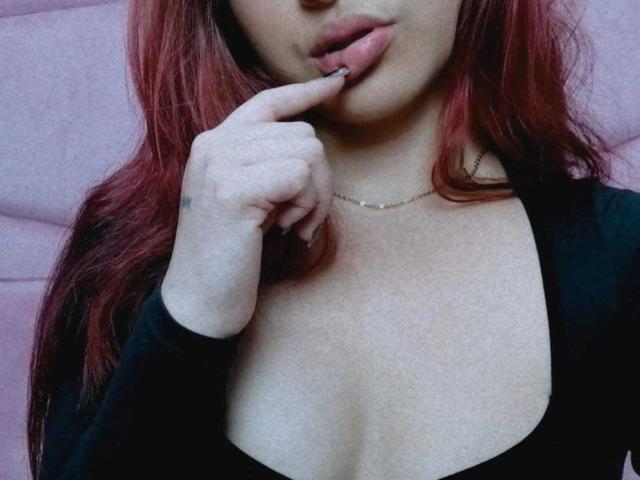 Samara, une jeune femme aux cheveux roux, portant un haut noir, avec un doigt dans la bouche, montrant ses seins, un décolleté subtil, une peau pâle, un collier en or, et les lèvres légèrement entrouvertes..