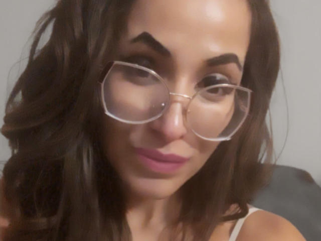 Adilay, avec ses cheveux bruns foncés et ondulés, portant des lunettes et du rouge à lèvres rose, regarde vers le bas avec un léger sourire. Ses seins sont petits, et elle a un visage mignon et séduisant..