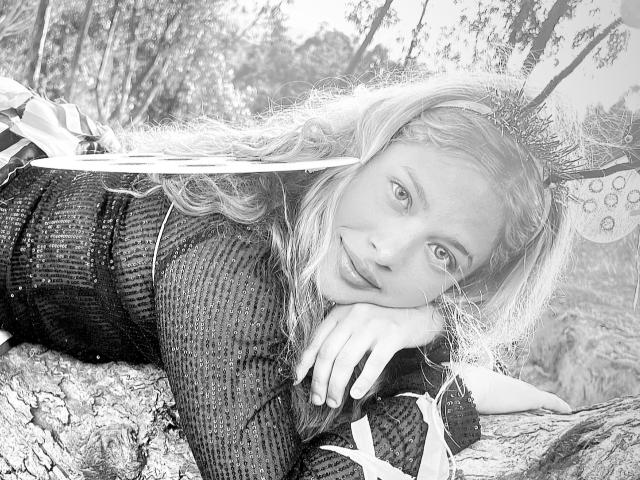 ShantalSummer with wavy blonde hair, wearing a ribbed black sweater, lies on a tree branch, eyes looking up, butterfly wings in her hair, hand resting on her chin.