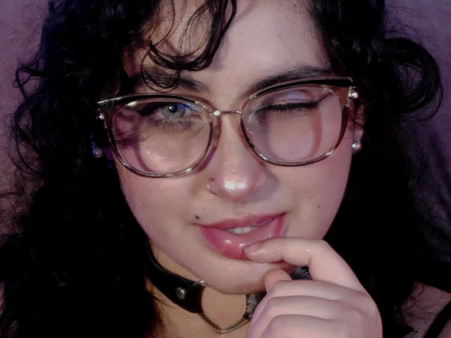 HannaCeleste with winking eye, wearing glasses, black curly hair, nose ring, choker, and touching her pink lips with finger. She's got a cute mole above her lip.