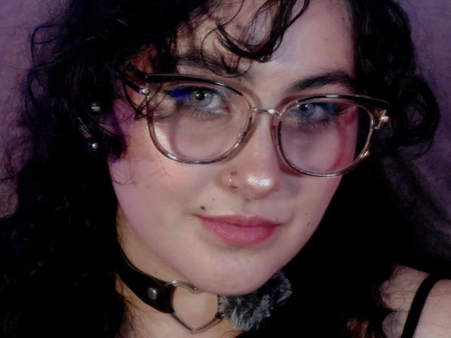 HannaCeleste with curly black hair, blue eyes, and glasses, wearing a black choker with a silver heart, nose ring, and pink lipstick, smiling slightly.