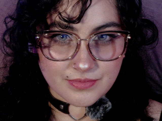 HannaCeleste with curly black hair, blue eyes, glasses, nose ring, and pink lipstick, wearing a black choker with fur trim, smiling softly. Her tits are not visible.