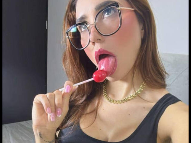 Bellasweeth, con gafas, lamiendo una piruleta roja, tiene lápiz labial rosa, esmalte de uñas rosa y un collar de cadena de oro, y lleva una blusa negra, con cabello castaño..