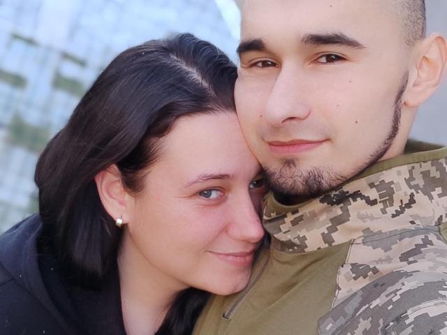 CharlotteAndJames, a woman with black hair and small earrings, is nuzzled against a man with a beard and camouflage jacket. She has blue eyes, and he has a slight smile.