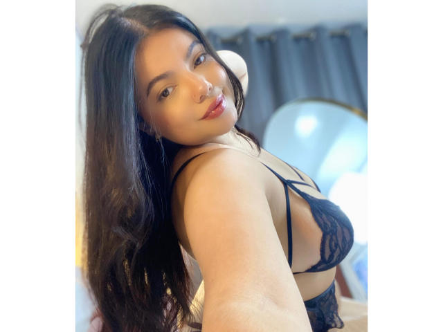 LaraJames with long black hair, wearing black lace lingerie, showing her tits in a selfie, smiling, looking at the camera, in a bright room with blue curtains.