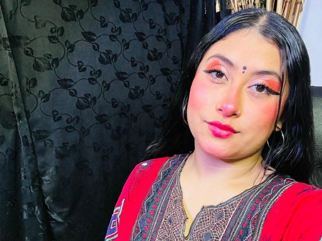 KalpanaAisha with dark hair, red eyeliner, pink blush, red lipstick, small black dot on forehead, hoop earrings, and red embroidered top, looking at camera with slight smile.