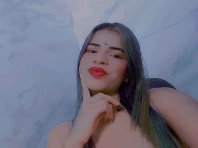 KairaDayamai with long dark hair, red lipstick, and a small forehead dot, touching her chin, smiling at the camera, showing her bare shoulders. Her hand has a green ring.