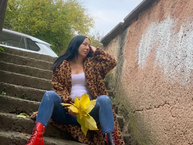 MirandaHott69 sits on worn, mossy stairs in a rough, textured setting. She has long black hair, a light brown leopard coat, white tank top, and blue jeans. Red lace-up boots contrast with the yellow leaves in her hand. Her right hand touches her hair while she looks slightly away. The weathered, pinkish wall on the right and green foliage in the background add texture and depth. Her relaxed, casual pose emphasizes the natural, everyday feel of the scene.