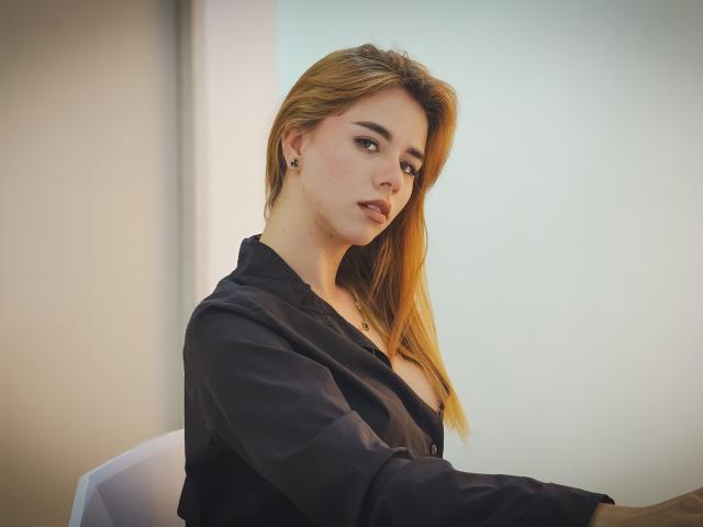 NoaBell with blonde hair, black shirt, and subtle makeup, sitting, showing her tits, looking serious, small black earrings, soft light, casual, relaxed, slightly open shirt, medium shot.