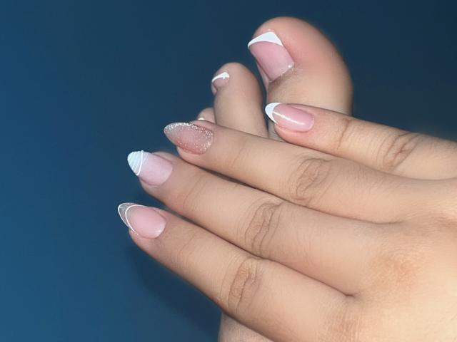 OlivaStellas' hands showing off her pink French manicure with white tips, sparkling glitter on the ring finger, and long, sharp nails against a dark blue background.