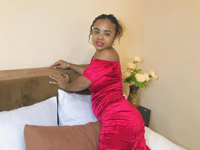JulianahMagik trägt ein rotes Samtkleid, lehnt sich an ein Bett mit weißen Kissen und braunen Polstern an, lächelt in die Kamera und zeigt ihr kleines Hinterteil und ihr lockiges Haar..