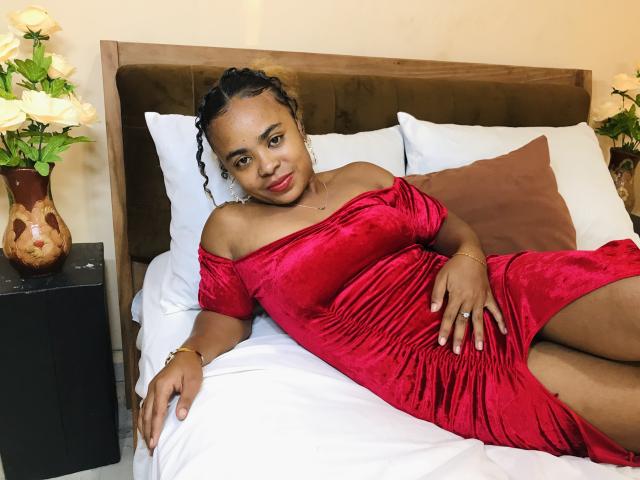 JulianahMagik vestindo um vestido vermelho de veludo, com um ombro de fora, deitada em uma cama, com a mão sobre sua região íntima, e um sorriso relaxado e sensual, cercada por travesseiros brancos e marrons..