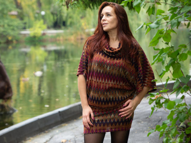 AlexisLovelyy with long red hair, wearing a colorful, zigzag-patterned, tunic dress, black tights, standing by a green, leafy, water-side, looking up with a slight smile, hands on hips.