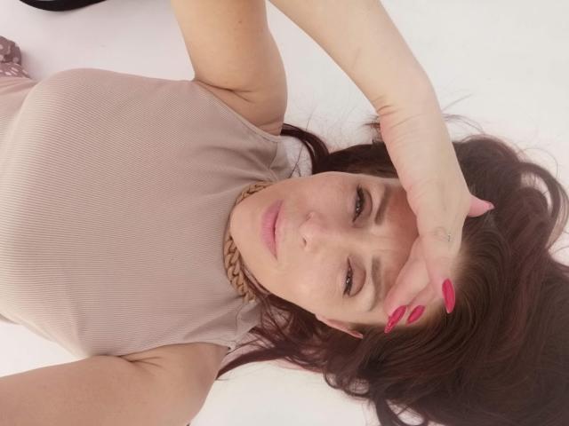 AlexisLovelyy lying on her back with dark brown hair, wearing a pink ribbed top, gold chain, and pink nails, covering her eyes with one hand.