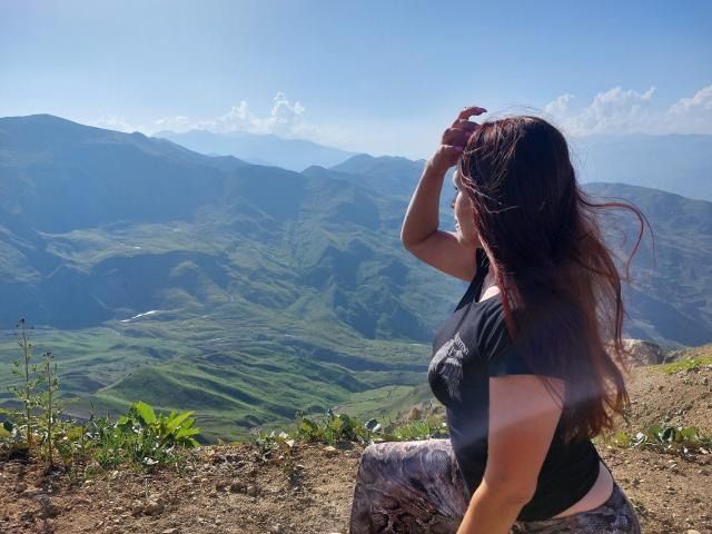 AlexisLovelyy with long brown hair, black top, and camo pants, sitting on a dirt ledge, hand in hair, looking at a green mountain landscape under a blue sky.
