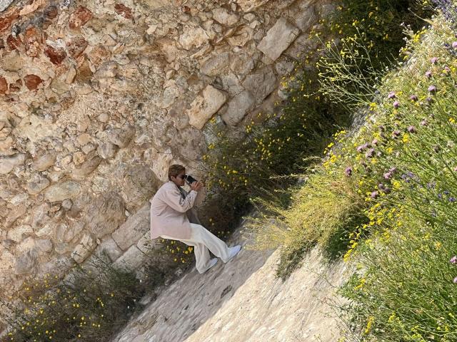 AnaRoa is crouching against a stone wall, wearing a pink coat and white pants, with a camera to her face, surrounded by greenery and yellow flowers.
