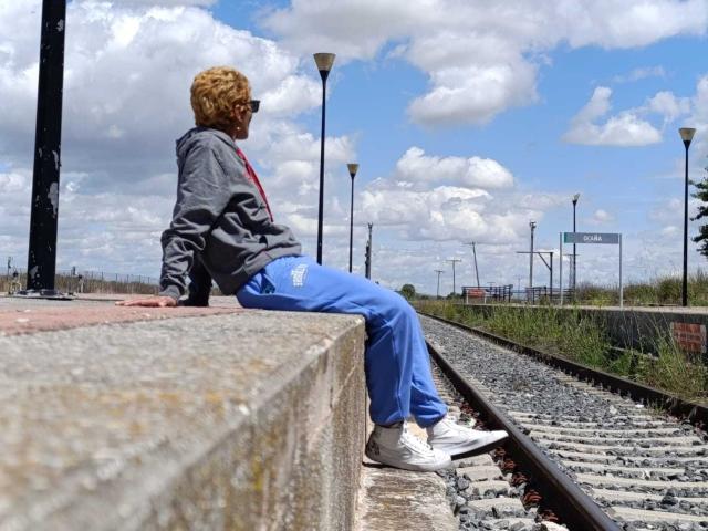 AnaRoa, with curly blonde hair and sunglasses, sits on a concrete platform in blue pants and white sneakers, looking away, in a cloudy sky with a train track.