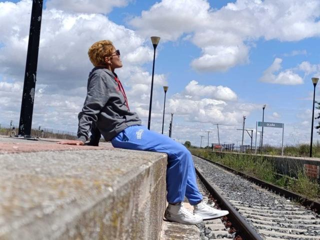 AnaRoa with curly blonde hair, wearing a grey hoodie, blue tracksuit, and white sneakers, sitting on a concrete ledge, looking to the side, with cloudy blue sky.