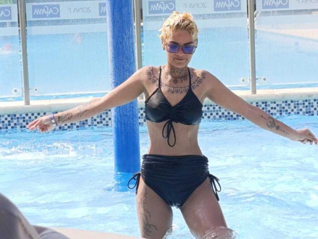 AnaRoa, con cabello rubio corto, tatuajes en los brazos y el pecho, vestida con un bikini negro, de pie en una piscina, con los brazos extendidos, agua salpicando, un poste azul detrás, gafas de sol moradas..