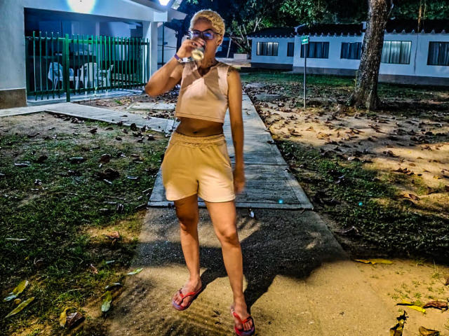 AnaRoa, avec des cheveux blonds courts, buvant dans une bouteille, portant un crop top rose, un short beige et des tongs rouges, se tient debout sur un chemin en béton la nuit..