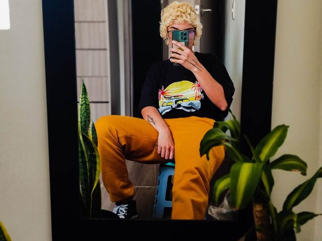 AnaRoa, avec ses cheveux blonds bouclés, un t-shirt noir, un motif coloré, un pantalon jaune, des baskets noires, prenant un selfie devant un miroir, assise sur un tabouret bleu, tenant un téléphone vert, avec des plantes en pot autour d'elle..