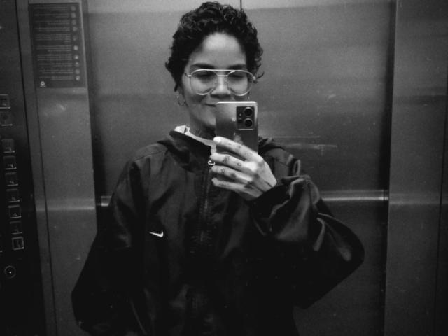 Ein Schwarz-Weiß-Selfie von AnaRoa mit lockigem Haar, Brille und einer dunklen Jacke, die lächelnd ein Handy hält und in einem Metallaufzug steht. Ihr Hintern ist nicht sichtbar..