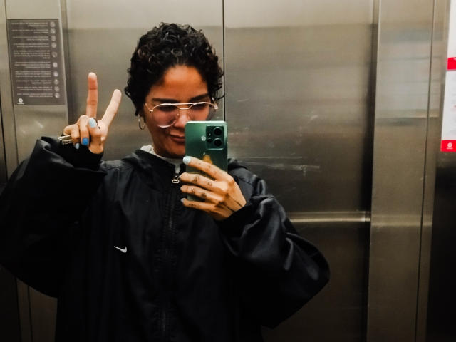AnaRoa, con cabello negro rizado, gafas y una chaqueta negra de Nike, sosteniendo un teléfono, haciendo un gesto de rock and roll con su mano derecha, dentro de un ascensor metálico..
