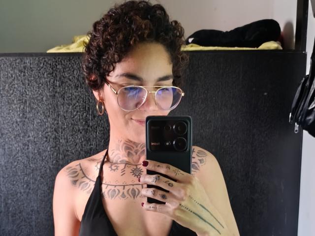 AnaRoa with curly brown hair, glasses, and black halter top, showing tattoos on neck and chest, holding phone, smiling, wearing gold hoop earrings, in selfie.
