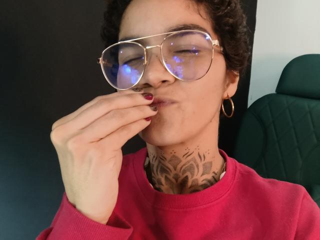 AnaRoa with curly hair, wearing gold glasses, red sweater, and hoop earrings, puffs out chest, showing neck tattoo, and red nail polish, with finger near lips.