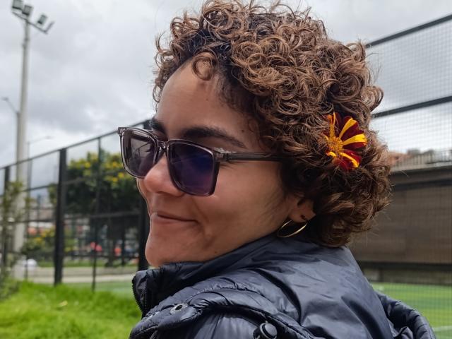 AnaRoa a les cheveux bruns bouclés avec une fleur rouge et jaune, elle porte des lunettes de soleil foncées et une veste noire matelassée, et elle sourit en tournant légèrement la tête..