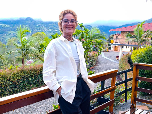 AnaRoa, com cabelos castanhos cacheados, óculos, camisa branca, calças pretas, sorrindo, com as mãos nos bolsos, em pé em uma grade de madeira, com muita vegetação, palmeiras e uma casa ao fundo..