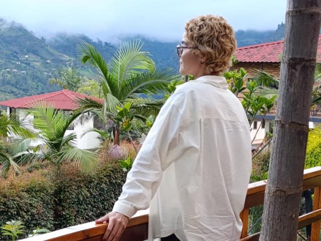 AnaRoa, con i capelli biondi ricci, che indossa occhiali e una camicia bianca, è in piedi su un balcone di legno, e guarda un paesaggio montuoso rigoglioso con palme e edifici con tetti rossi..