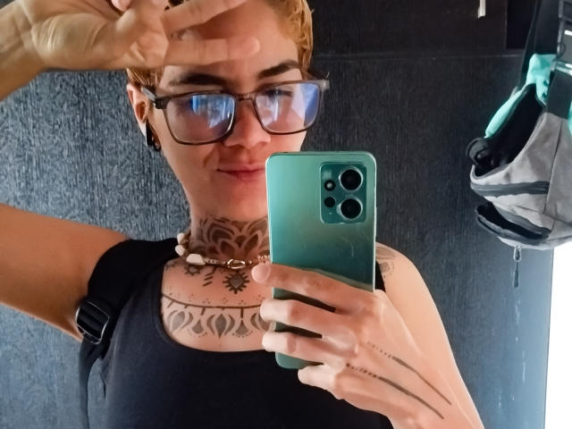 AnaRoa, con cabello rojo corto, gafas, camiseta negra, tatuajes en el cuello y el pecho, sosteniendo un teléfono verde, haciendo el gesto de la paz, sonriendo, con una correa de mochila negra visible..