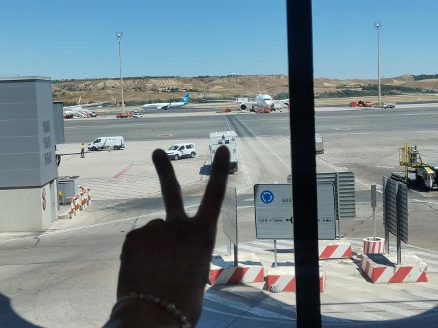 La mano de AnaRoa hace un gesto de la paz, con el dedo levantado, en primer plano. En el fondo, se ve un avión en la pista, varios vehículos y un cielo azul despejado..