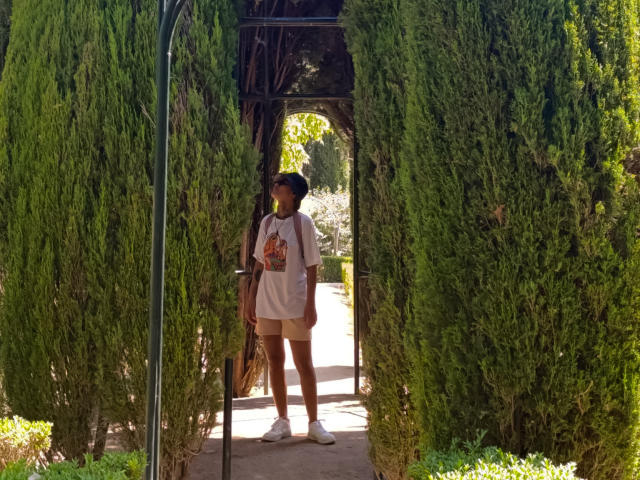 AnaRoa si trova tra alti cespugli verdi, indossando una maglietta bianca con un motivo colorato, pantaloncini beige e scarpe da ginnastica bianche, sotto un arco di legno scuro illuminato dal sole..