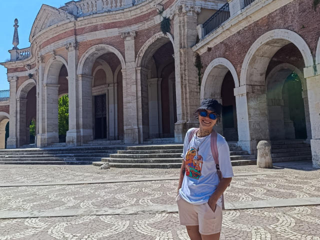 AnaRoa è in piedi di fronte a un edificio in mattoni, indossa una maglietta blu tie-dye con una grafica colorata, pantaloncini beige, un berretto nero e occhiali da sole, sorridendo con le mani in tasca..